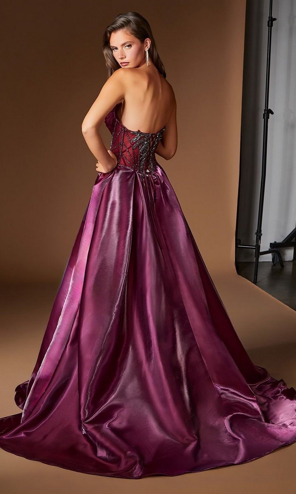 A woman stands in a studio, looking over her shoulder, wearing the Ladivine CDS568 long prom dressโa strapless, floor-length couture gown in shiny burgundy featuring a fitted corset back and a full organza skirt.